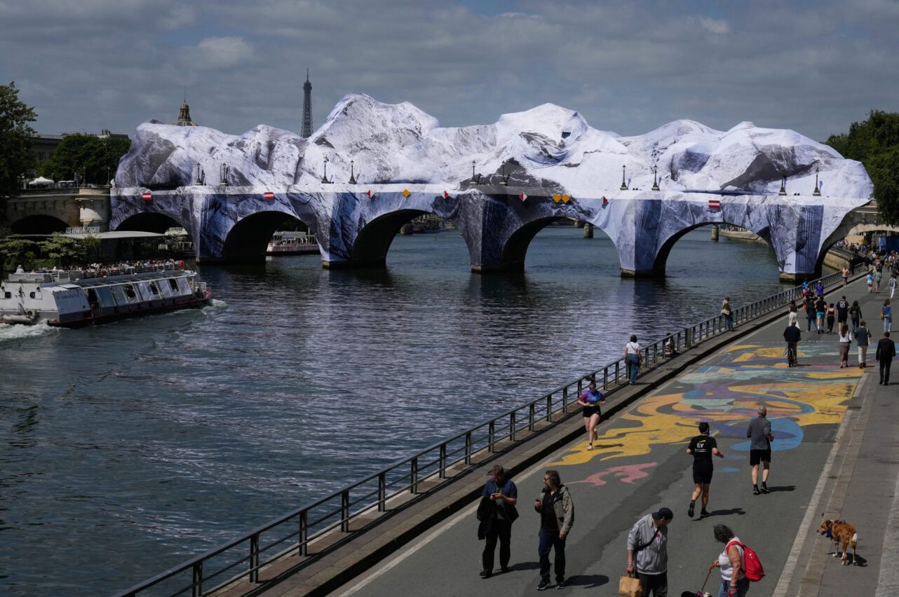 Brücke mit weißer Hülle, darunter fließt ein Fluss, daneben Menschen und Boote.