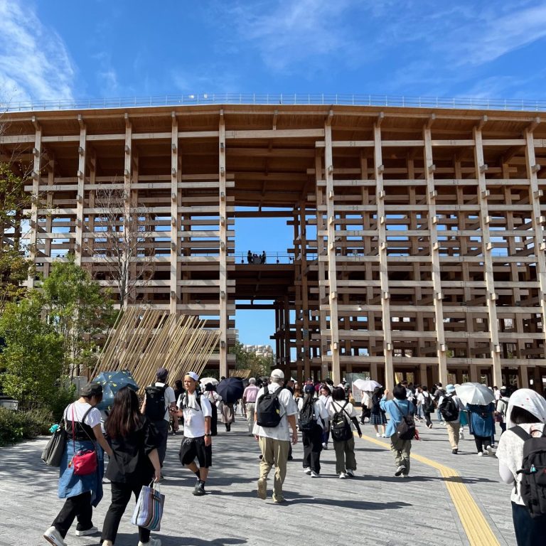 Holzgebäude mit vielen Menschen davor, blauer Himmel mit wenigen Wolken.
