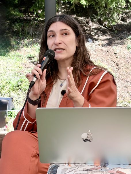eine Frau sitzend spricht mit Mikro - im Vordergrund ein aufgeklappter Laptop