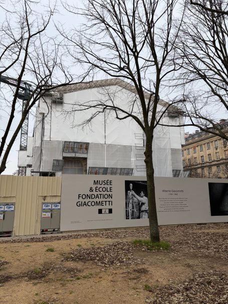 Baugerüst um ein unvollendetes Gebäude, umgeben von Baustellenzäunen und Bäumen.