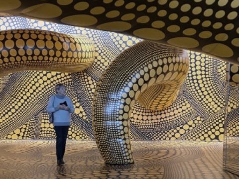 Eine Person steht in einem Raum mit geschwungenen Wänden und goldenen Punkten.