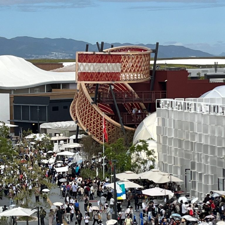 Große Menschenmenge vor modernem architektonischen Gebäude des österr Pavillions mit geschwungenen Elementen und integrierter Fahne.