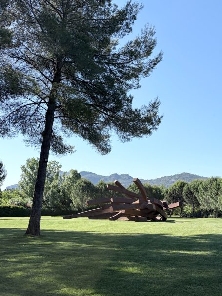 Skulptur im Freien, umgeben von Bäumen und Wiesen, im Hintergrund Berge sichtbar.