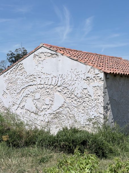 Wand mit Reliefkunst und Augenmotiv, umgeben von grünem Gras und blauem Himmel.