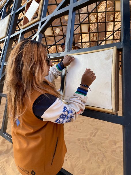 Frau mit langen Haaren schreibt auf einem weißen Blatt an einer Wand aus Gitter.