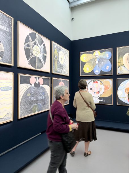 Eine Kunstgalerie mit mehreren Bildern an Wänden, Besucher betrachten die Werke.