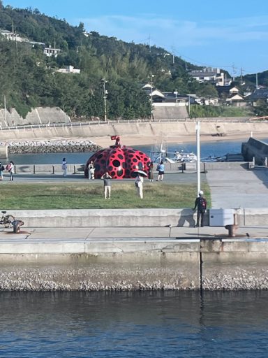 Eine rote Skulptur auf einer Wiese am Hafen, umgeben von Personen und Bäumen.