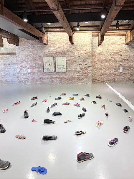 Kreative Installation von Schuhen auf einem hellen Boden in einem Raum mit Backsteinmauer.