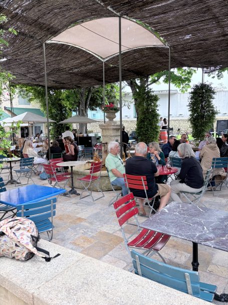 Gemütliches Café im Freien mit Gästen an Tischen unter einem Sonnendach.