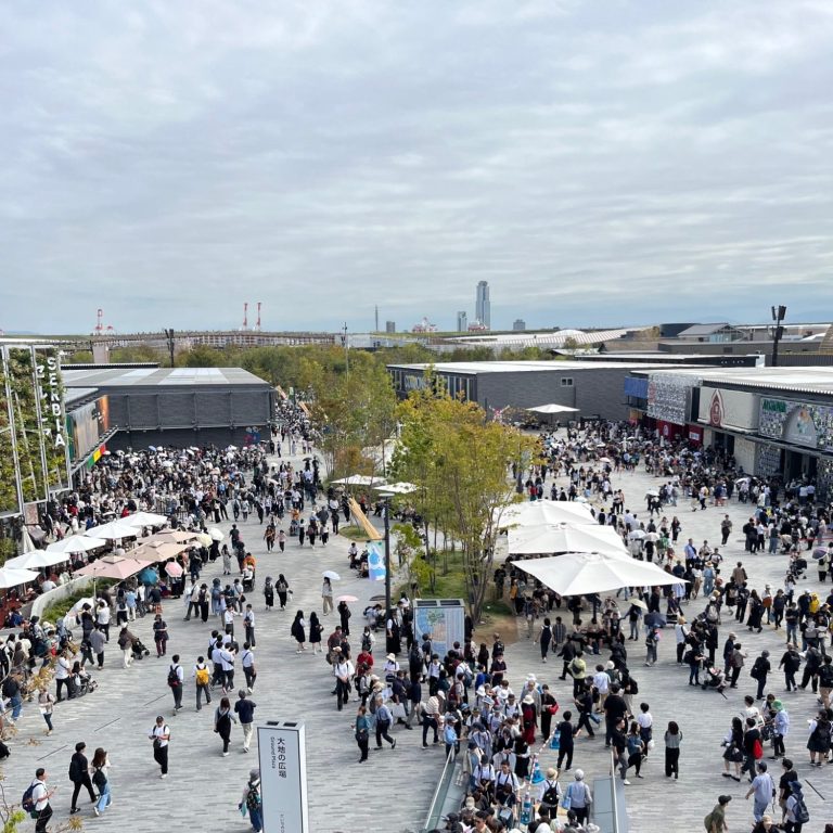 Belebter Außenbereich mit Menschenmengen, Ständen und cloudy Himmel im Hintergrund.