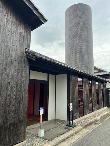 Moderner zylindrischer Turm neben traditionellem Holzgebäude in einer urbanen Umgebung.