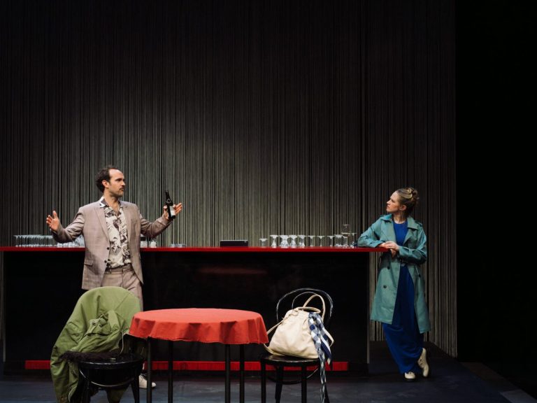 Zwei Schauspieler stehen in einer Theaterszene, vor einem Tisch und einer Bar.