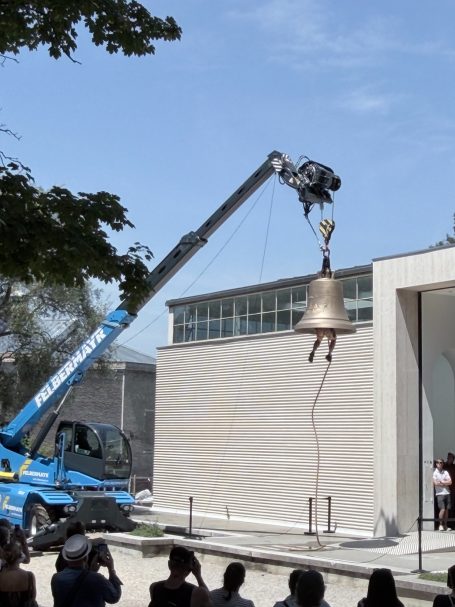 Eine große Glocke wird mit einem Kran aufgestellt, Zuschauer beobachten gespannt.